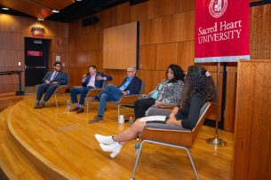 On a large wooden stage, four trustees and a student sit in wooden chairs facing an audience for a Q&A-style discussion.